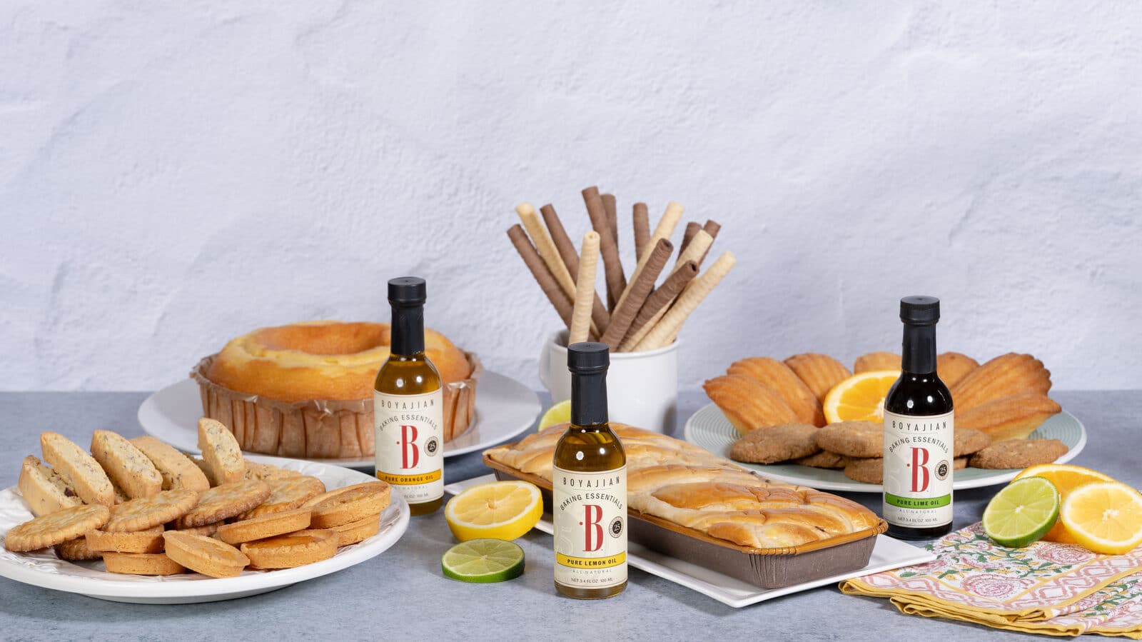 A collection of Boyajian Pure Orange, Lemon, and Lime Oils displayed with various baked goods including a bundt cake, biscotti, madeleines, wafer rolls, and sliced citrus fruits.