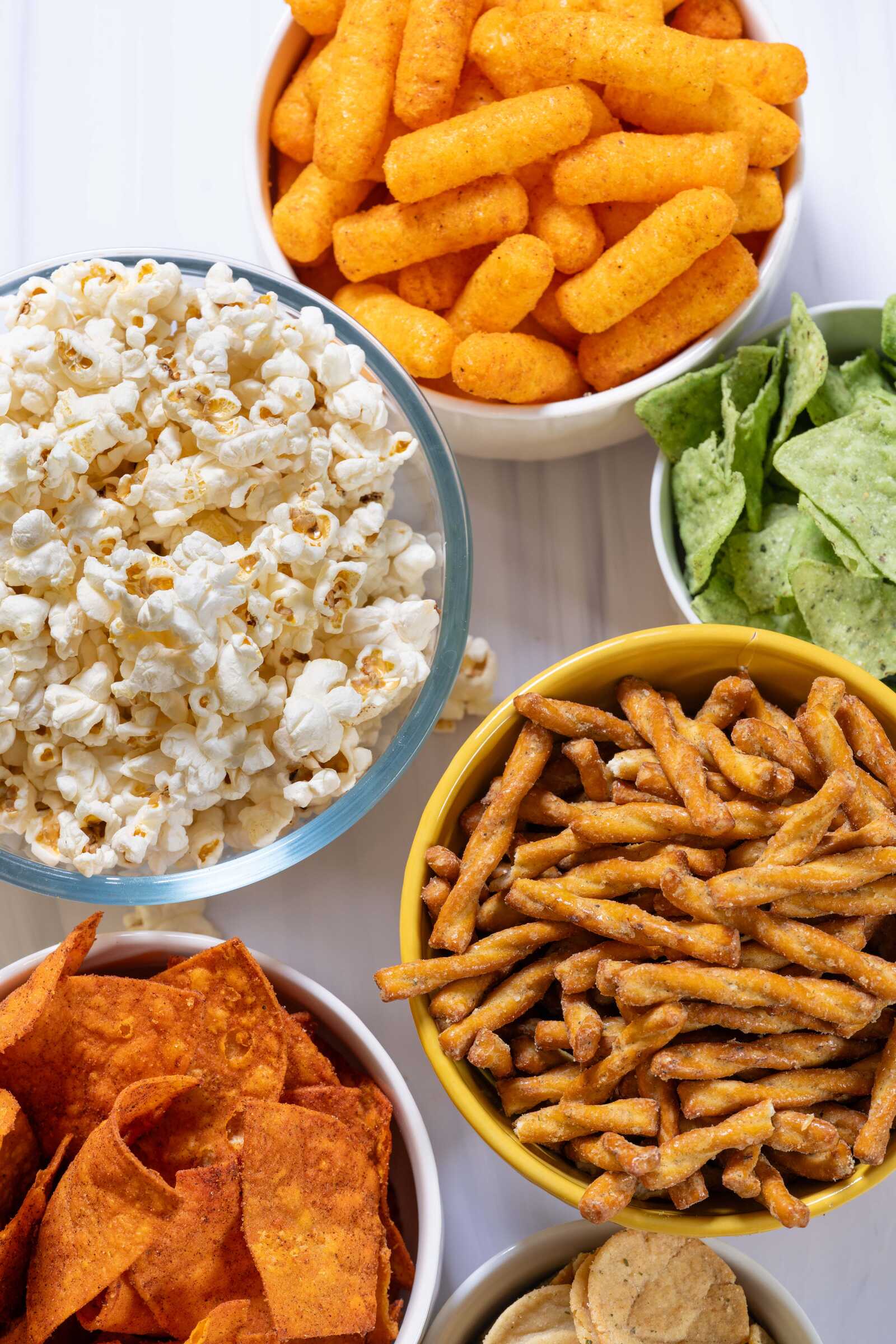 Six bowls of snacks: popcorn, orange cheese puffs, green tortilla chips, twisted pretzel sticks, orange tortilla chips, and round cracker chips, on a white background.