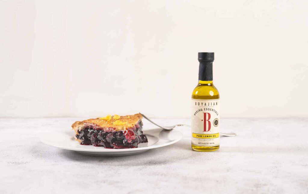 A slice of berry pie with lemon zest on a white plate next to a bottle of Boyajian Pure Lemon Oil.
