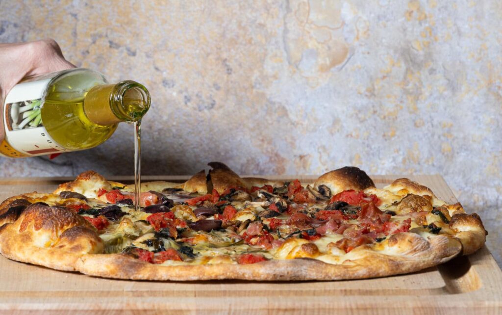 A hand pours olive oil from a bottle onto a round pizza with cheese, tomatoes, mushrooms, and olives, resting on a wooden board.