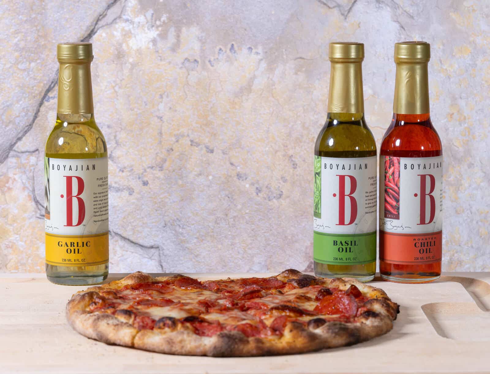 Three bottles of Boyajian oils (Garlic, Basil, Roasted Chili) stand behind a pepperoni pizza on a wooden board.