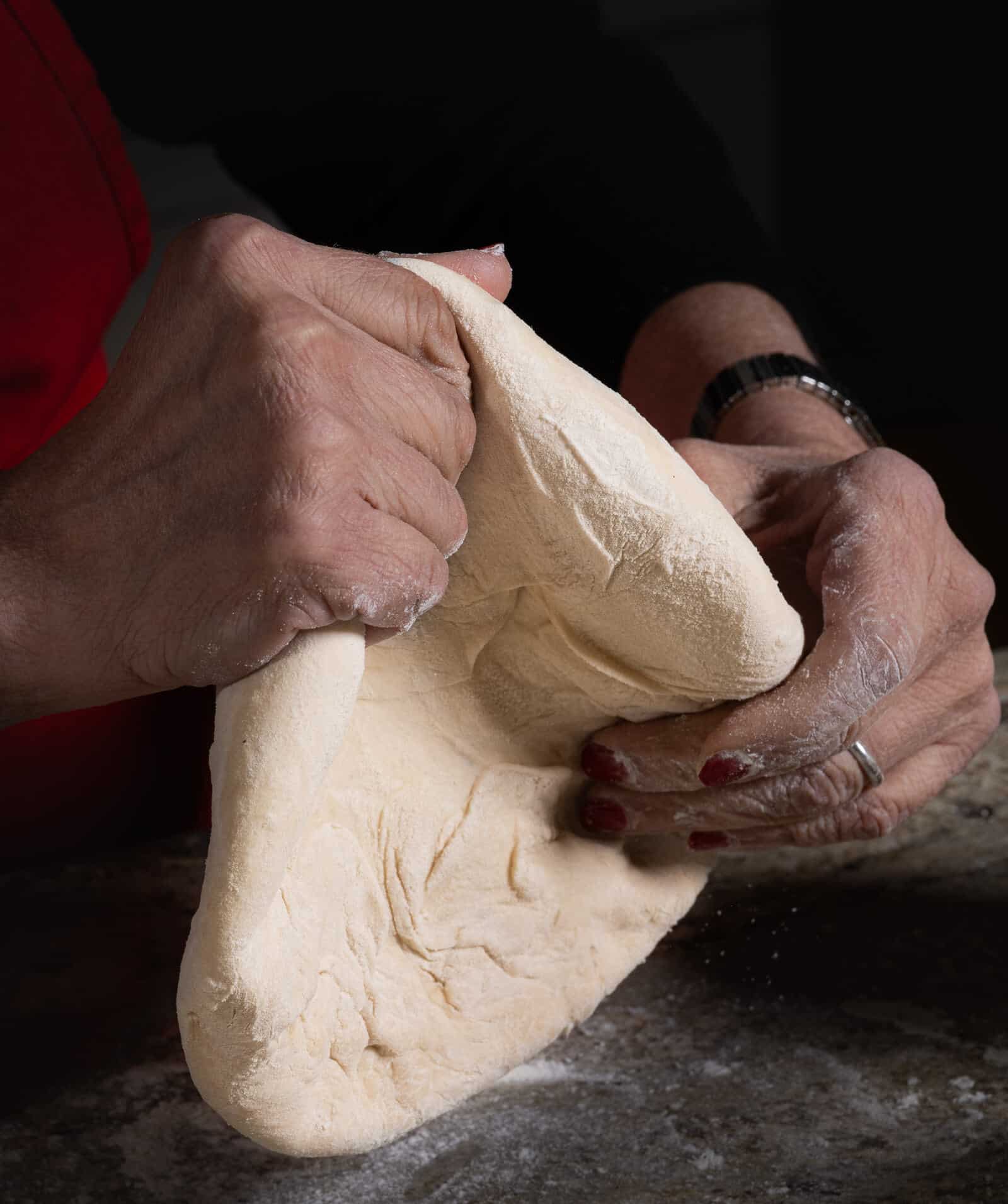 Two flour-dusted hands, one with red nail polish and a ring, are kneading a large piece of light-colored dough on a dark, floured surface.