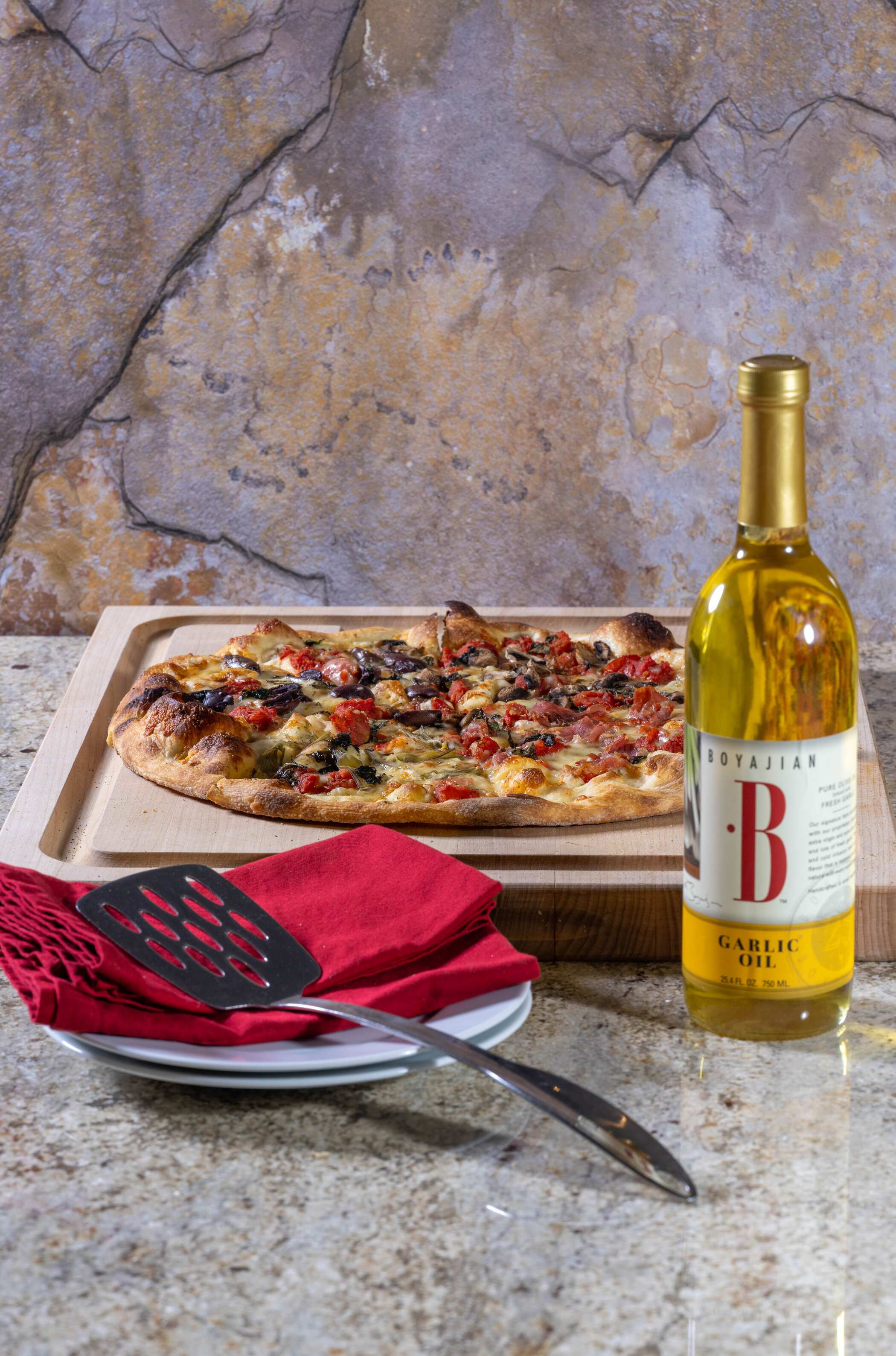 A pizza with tomatoes, olives, mushrooms, and ham on a wooden board, next to a bottle of garlic oil, plates, and a spatula on a red napkin.