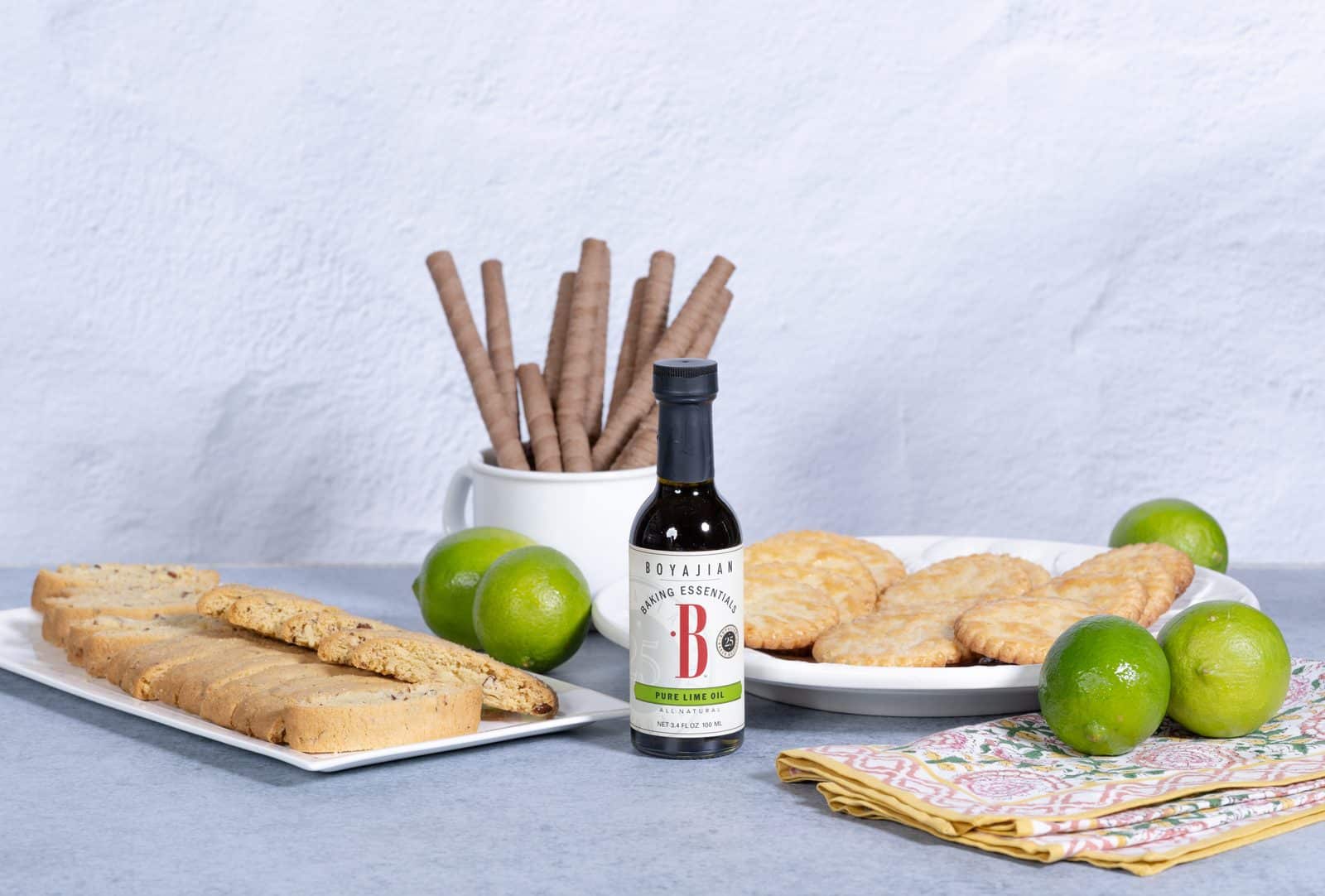A bottle of Boyajian Pure Lime Oil surrounded by biscotti, small pastries, fresh limes, and chocolate wafer rolls on a gray surface.