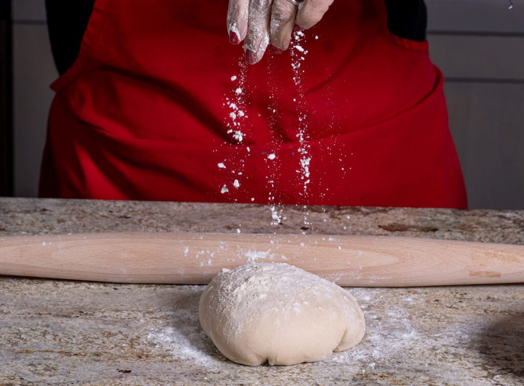 A person in a red apron sprinkles flour onto a ball of dough on a granite counter, with a rolling pin nearby.