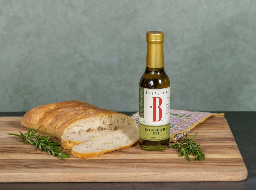 A wooden cutting board holds a loaf of bread with two slices, a bottle of Boyajian Rosemary Oil, and fresh rosemary sprigs. A colorful napkin is behind the bottle.