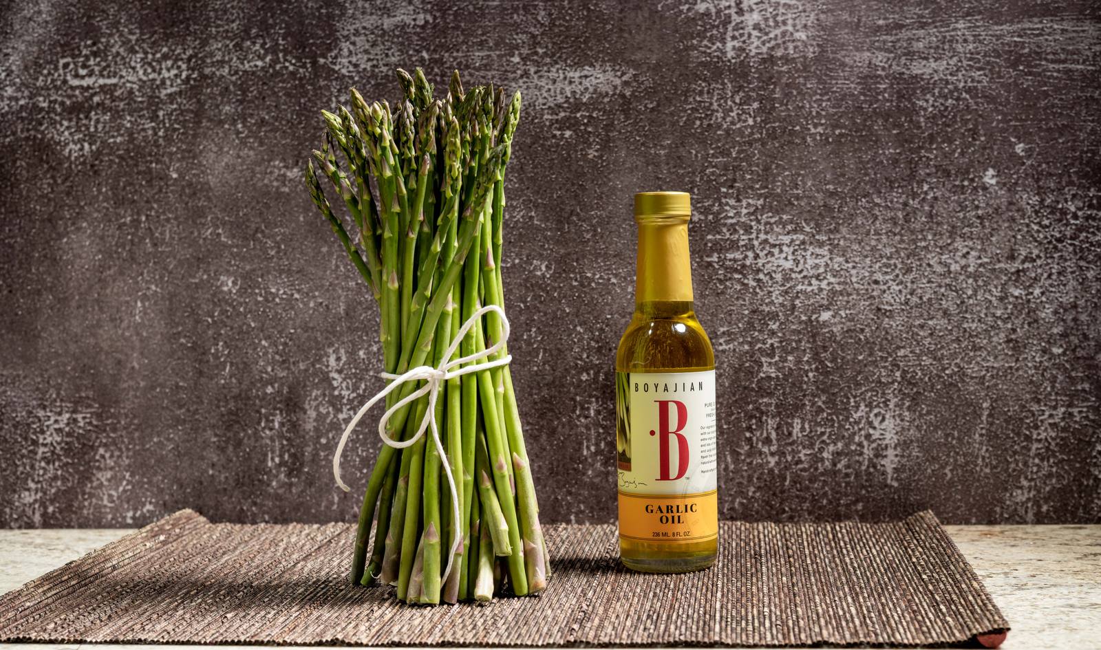 A bundle of green asparagus tied with white twine next to a bottle of Boyajian Garlic Oil, on a dark mat.