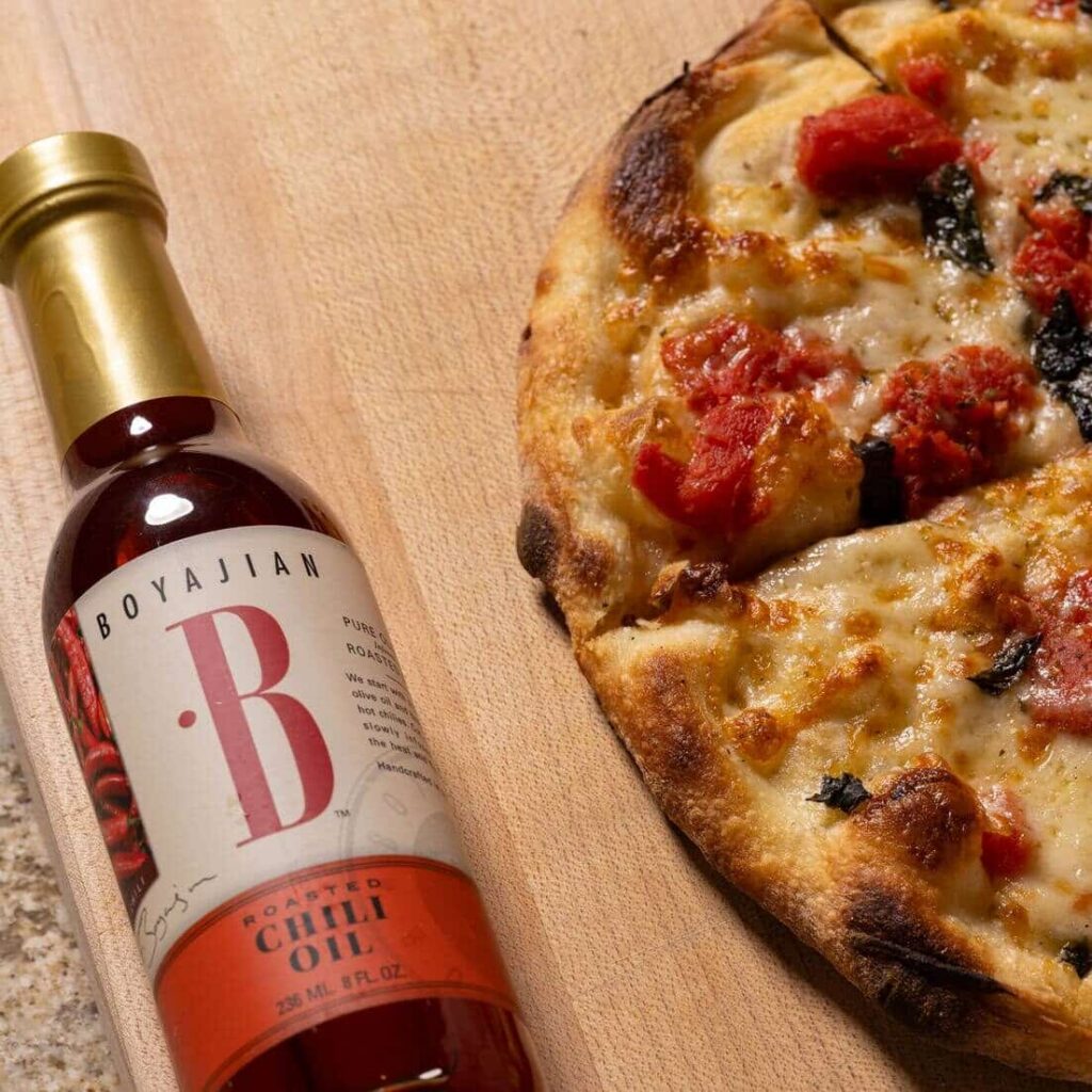 A bottle of Boyajian Roasted Chili Oil next to a pizza with cheese, tomato, and basil on a wooden board.