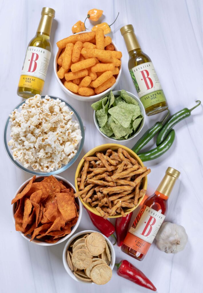 An overhead view of various snacks including popcorn, cheese puffs, green chips, pretzel sticks, and red chips in bowls, alongside three bottles of Boyajian flavored oils (Garlic, Jalapeño, Roasted Chili), and fresh peppers and garlic, all on a white marble surface.