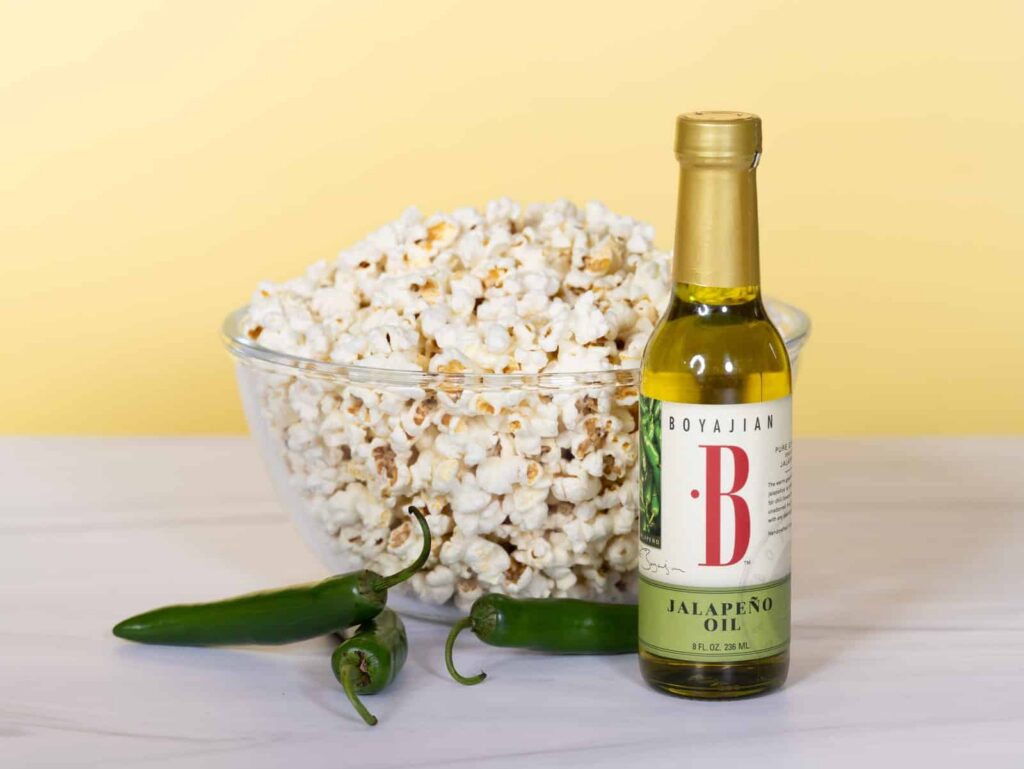 A bowl of popcorn, a bottle of Boyajian Jalapeño Oil, and three green jalapeño peppers on a light surface with a yellow background.