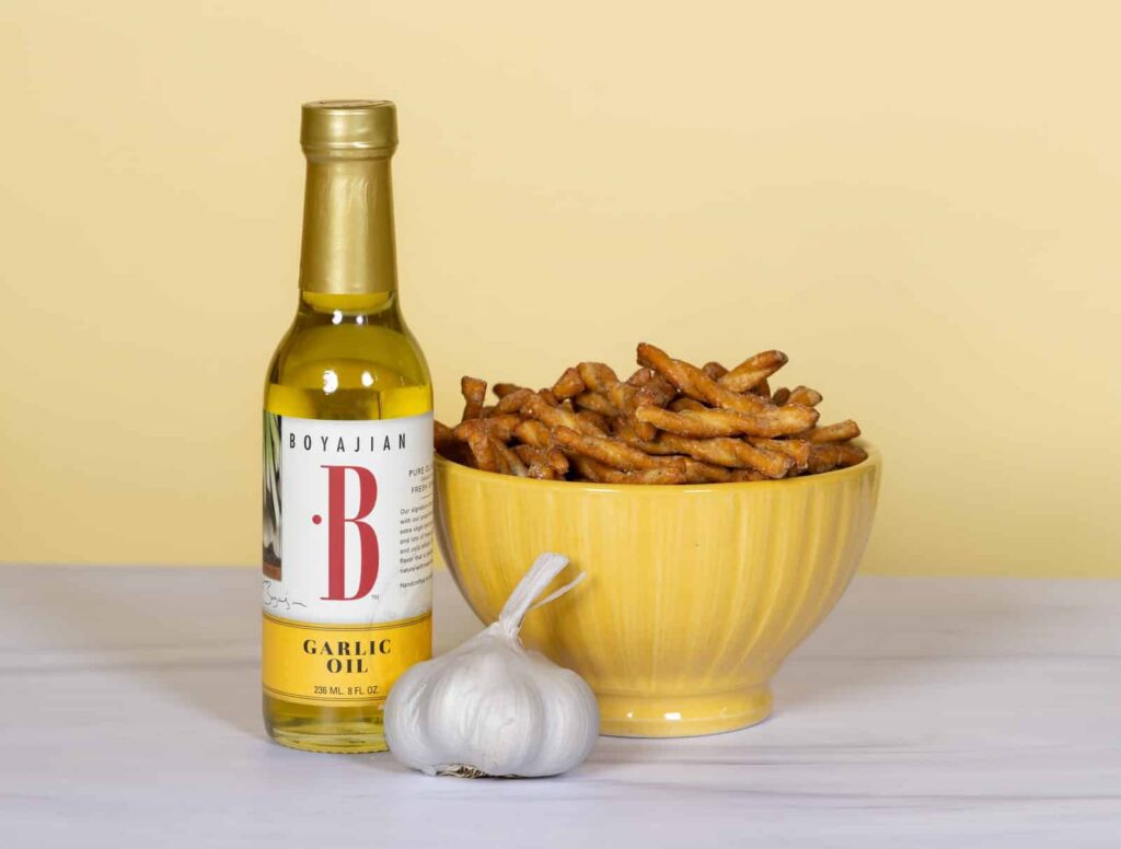 A bottle of Boyajian Garlic Oil, a yellow bowl of seasoned pretzel twists, and a garlic bulb on a light background.