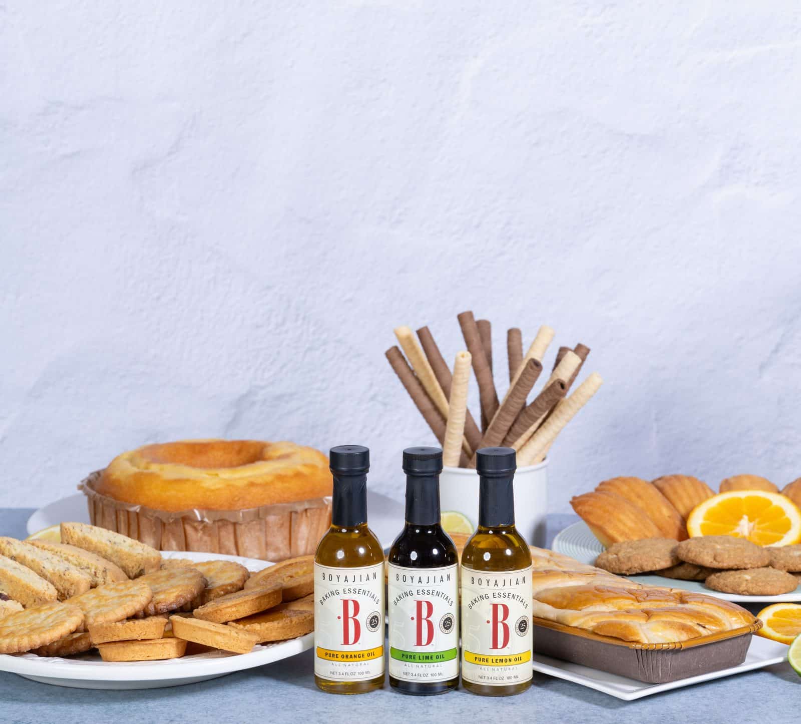 Three bottles of Boyajian Pure Orange, Lime, and Lemon Baking Oils are surrounded by a variety of baked goods including a bundt cake, cookies, madeleines, and wafer rolls.