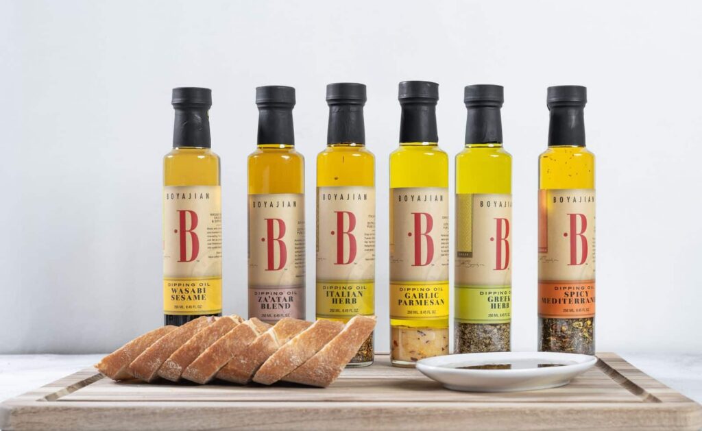 Six bottles of Boyajian dipping oils in various flavors, including Wasabi Sesame, Za'atar Blend, Italian Herb, Garlic Parmesan, Greek Herb, and Spicy Mediterranean, displayed on a wooden cutting board with sliced bread and a small dish of oil.