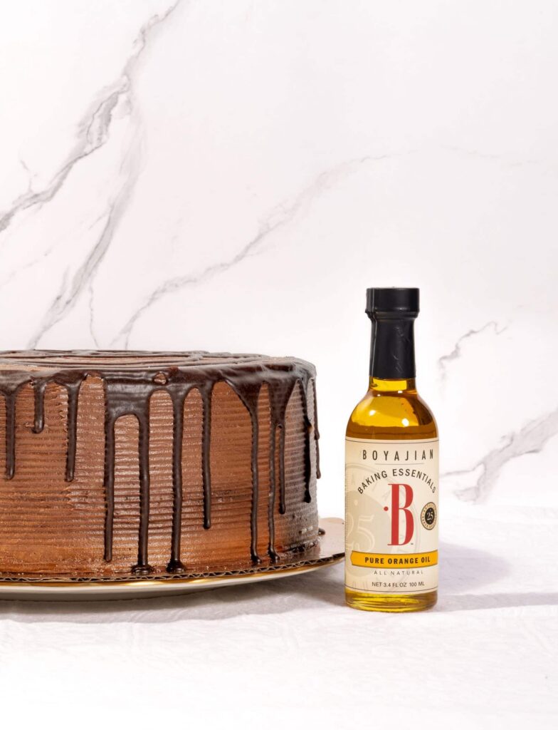 A chocolate cake with ganache drips next to a bottle of Boyajian Pure Orange Oil on a white marble background.